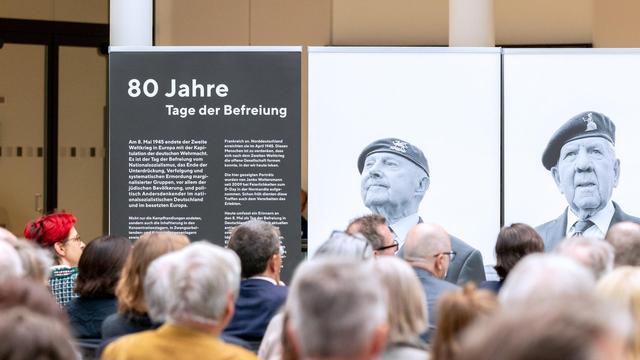 Menschenmenge von hinten, sie blicken in Richtung eines Aufstellers mit der Aufschrift '80 Jahre Tage der Befreiung'. Daneben auf Aufstellern zwei schwarz-weiße Portraits von Veteranen.