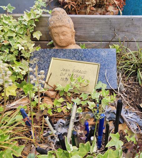 Une plaque de couleur dorée avec écrit : Jean Giraud
Dit Moebius, 
1938 - 2012
La tête d'une statuette qui soutient cette plaque.
Au premier plan, des rotrings plantés dans une végétation spontanée faite d'herbes et de lierres.