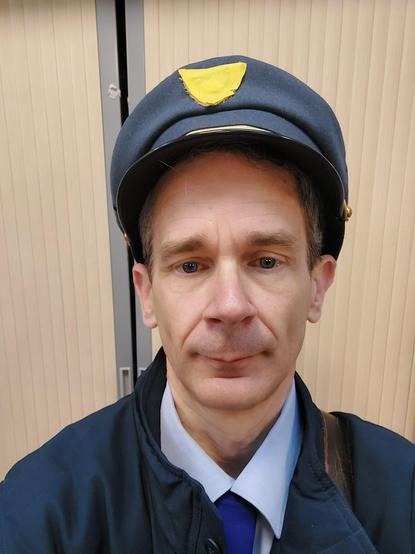 Selfie en postier casquette bleue comme couvre chef, cravate, chemise, veste bleue et sacoche de cuir en bandoulière.