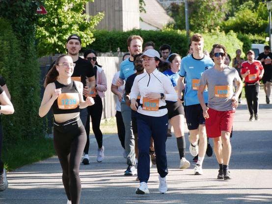 A group of runners, led by several people with visible bib numbers, runs along a sunny street. The mood is focused and energetic.