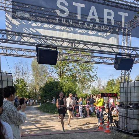 Two runners cross the finish line side by side under a banner that reads “START TUM Campuslauf,” surrounded by cheering spectators. A digital clock displays the race time.