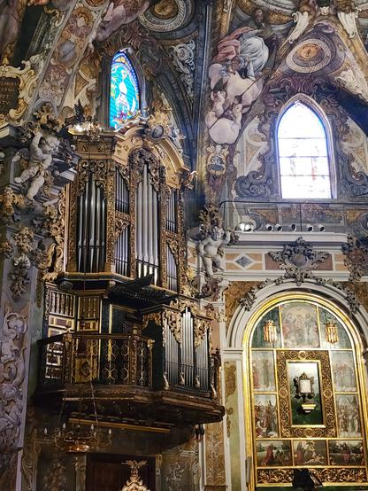 To the left, large church organ high on the wall with dark metallic pipes and gold-colored Rococo metalwork covering their entire frame. Surrounding it and on the wall directly facing viewer are frescoes resembling those of the Sistine Chapel and two small stained glass windows.
