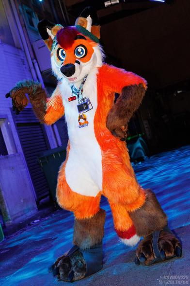 Colto Fox (tall orange, white, brown and red fox fursuiter) in full fursuit standing in an alley with a water textured blue light illuminating his surroundings. He has his left hand on his hip and his right hand up with his wrist folding his hand downward.