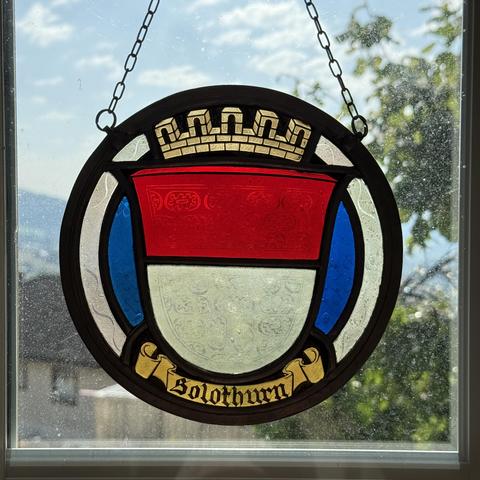 A view from our kitchen window. It shows a leadlighting of the emblem of Solothurn. The emblem shows the Solothurn colors white in the lower part and red in the upper part. On top of it there is a castel wall, below is a scroll with the inscription "Solothurn". To the left and right, there are blue areas, symbolizing the river Aare.

The whole emblem is made of colorful glass which is held together with lead frames. All this is surrounded be a round frame. It's very colorful if the sub shines through it.