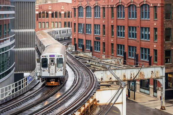 A brown line CTA train moves between buildings in Chicago. Multiple buildings are on the outside, and the train is in the middle following an S curve.