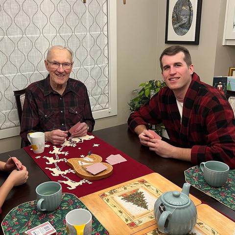 Photo of an older man (my grandfather) & younger man (me) holding cards sitting at a table lain with Christmas-themed decorations and a cribbage board.