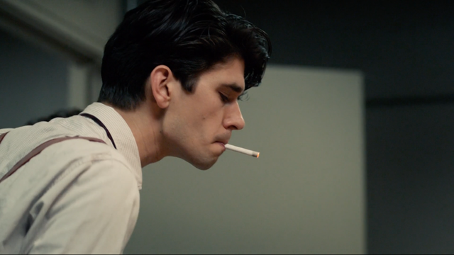 This is a photo of Freddie Lyon from The Hour. He's in the office, wearing a white shirt with a cigarette hanging out of his mouth, and he's leaning forward to look at something off camera.