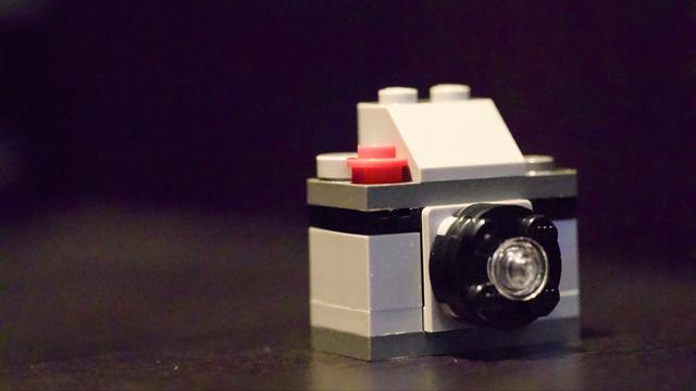 A tiny LEGO camera on a black shelf. The image is very noisy since it was shot at ISO 25600 (per the challenge).