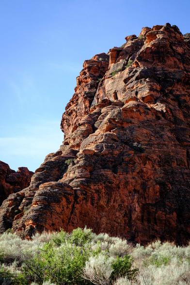 A large rocky formation with reddish-brown layers rises above sparse green and grayish shrubs, set against a clear sky.