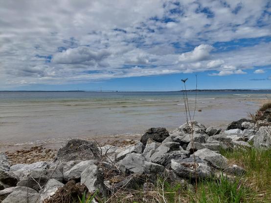 A view from the shore of Macinac Island. The sky is mostly cloudy, with a lot of blue towards the horizon and white clouds above. The Macinac bridge and mainland are visible off in the distance (the lower peninsula on the left and upper peninsula on the right). In the foreground, there is a rocky beach, then larger rocks and some grasses.