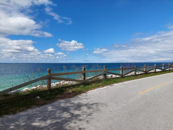 The deep blue of Lake Huron meets a partly cloudy sky in the background. In the foreground is the cycling and pedestrian path that encircles the island and a wooden fence.