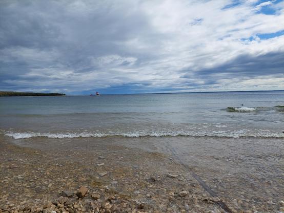 Waves break along the rocky shore. On the horizon are a lighthouse and another island with a mostly cloudy sky above.