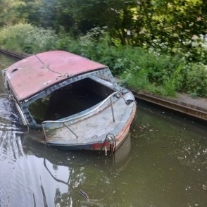 A sunken pleasure boat, missing windows, full of water, sits at an awkward angle on the bottom of a watery canal. While one gunnel is just under water, perhaps a third to half the boat is above the water level.

The boat has a faded red roof, and the foredeck might be a faded blue.

The boat, though sunken, is tied along the canal's off side (not the towpath side), where there is plenty of green scrub, bushes, and trees.

The canal bank is hardened with a steel rail. The water is a pale grey-green.