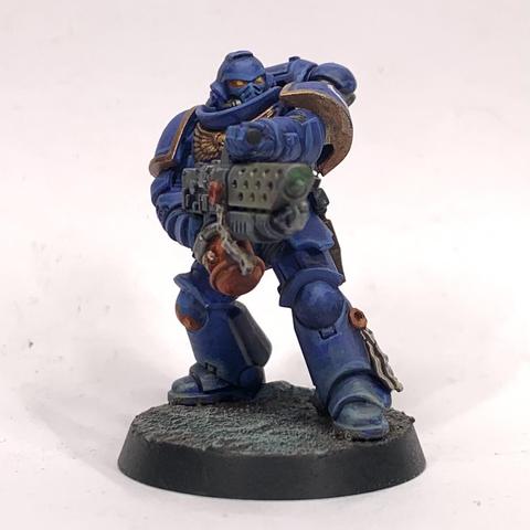 A Space Marine with a flame thrower. They wear bulky blue armor and aim their heat-bloomed weapon to their left.