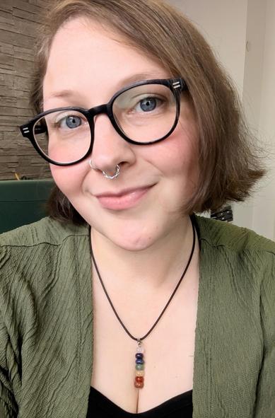 Ein Selfie von mir auf der Arbeit. Ich trage eine. Schwarze Brille, eine grüne Strickjacke und ein schwarzes Top. Eine Kette mit einem bunten Anhänger aus Steinen.