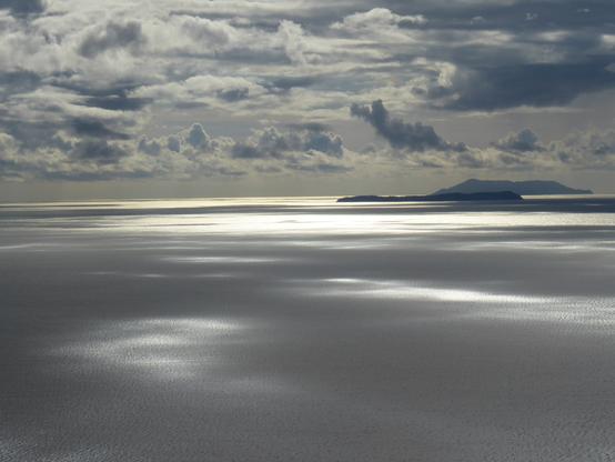 View from a vantage point west into the sun over the Ionian Sea. Mottled cloud sends reflections over the flat sea. A couple of islands lie on the horizon.