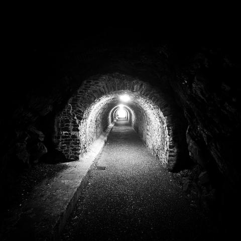A black and white image of a tunnel . Tue outside of tue picture is pitch black, with the tunnel lit in the middle of the picture, looking down the empty tunnel.