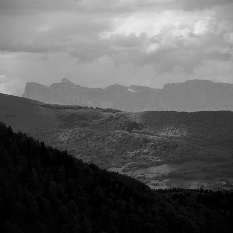 Photo noir et blanc d'un paysage avec plusieurs plans.
D'abord des collines boisées dans l'ombre.
Puis Des collines semi-boisées parsemées de taches de soleil.
Puis de hautes falaises avec quelques névés.
Enfin de ciel chargé de nuages bourgeonnants.