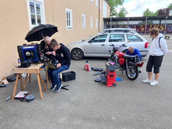 3 Personen arbeiten an verschiedenen Reparaturobjekten auf einem Parkplatz: Ein Stromerzeuger und ein Motorrad der Marke Simson