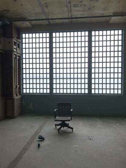 An older-style office chair is shown in the middle of a bare floor in a dimly lit room. The back wall is dominated by an opaque-glass window divided into numerous subpanels.