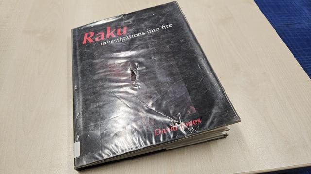 A book on a table top, it has a dark/black cover wrapped in clear film, in red at the top the word: Raku

In white under the title: investigations into fire

And at the bottom: David Jones