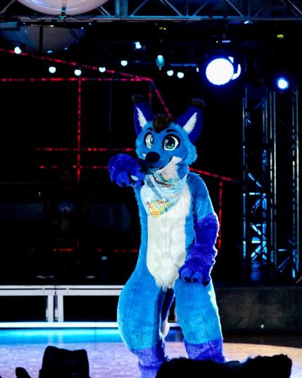 A blue fox fursuiter pointing at the audience while dancing in a darkened room. The fursuiter is wearing a bandanna from Furnal Equinox