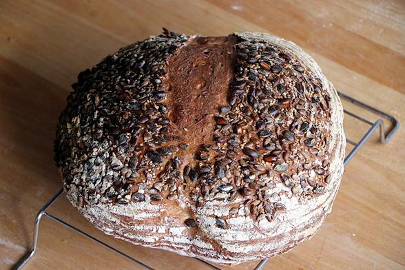 A large round loaf, heavily seeded with mixed seeds everywhere on top except where the score has expanded with the oven spring. The sides just showing a little of the flour markings typical of final proofing in a banneton.