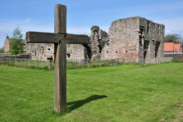 The ruins of Balmerino Abbey. The image shows a broad grassy area on the far side of which is low wooden fencing and partial stone ruins, with the most substantial remains on the right. In the left foreground is a large wooden cross planted in the ground. The sky is largely blue.