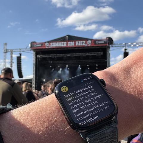 Lautstärke-Warnung auf einer Apple Watch. Im Hintergrund sind Menschen und eine Bühne zu sehen, ein Banner besagt „Sommer am Kiez“