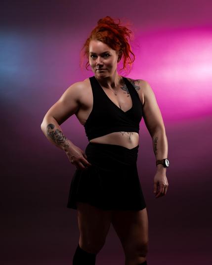 The image features a person with red hair posing confidently against a backdrop with pink and blue lighting. The individual is wearing a black outfit, and you can see tattoos on their arms. The setting gives an artistic and vibrant feel.