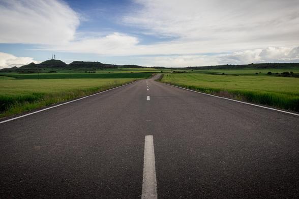 Photograph showing a road from a low angle. Green fields are visible on the sides, and a blue sky with several white clouds is visible in the upper third.