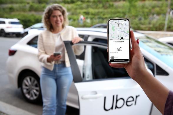 Eine Person hält ein iPhone mit Uber-App in die Kamera. Im Hintergrund eine Uber-Fahrerin an der Tür eines weißen Uber-Autos. Foto (c) Uber.