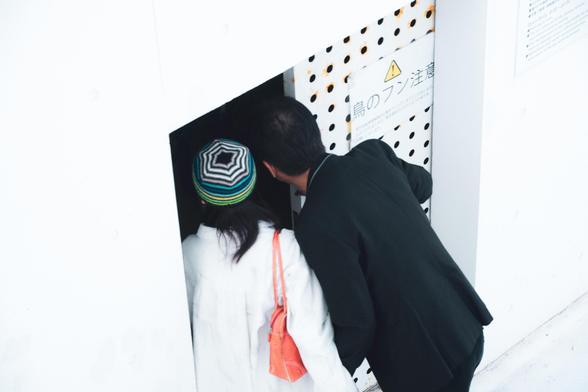 The image shows two people peering into an opening in a white wall. One person is wearing a multicolored hat and has a red bag, while the other is dressed in dark clothing. There’s a sign with Japanese text next to them, which includes a triangular warning symbol.
