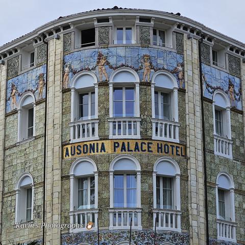 Jugendstilfassade des Ausinia Palace Hotel am Lido, Venedig, verziert mit Blumengirlanden tragenden Putten auf hellblaumem, gefliesten Hintergrund. Weiß gerahmte Fenster mit Rundbogen, der Hotelname ist mit goldfarbenen Fliesen hinterlegt.