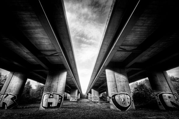 Low angle, black and white photograph of a bridge with symmetrical concrete supports and graffiti, leading to a bright opening in the sky.