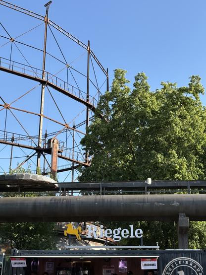Unter blauem Himmel im Vordergrund ein Getränkestand mit „Riegele“ Leuchtschrift, dahinter ein großer Baum rechts und das verrostete Gestänge eines Teleskopgasbehälters, alles im Anschnitt. Eine typische Mischung im Sommer im Alten Gaswerk Augsburg.