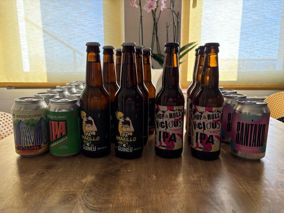 Bottles and cans of craft beer only it ch en table. There are orchids in the background in between the two windows.