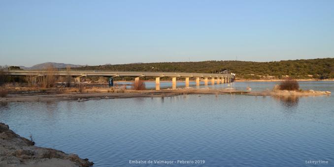 Fotografía del largo puente de la carretera M-505 que cruza el Embalse Valmayor tomada en Febrero de 2019.
El nivel de agua está muy bajo, hay columnas del puente que están sin cubrir de agua.
Se ven algunos arbustos secos. 
La foto está tomada desde el lado que va de El Escorial hacia Galapagar.