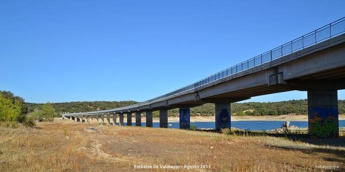 Fotografía del largo puente de la carretera M-505 que cruza el Embalse Valmayor tomada en Agosto de 2015.
El nivel de agua está muy bajo, hay columnas del puente que están sin cubrir de agua. En algunas zonas se puede pasar por debajo del puente porque no hay agua. El suelo está muy seco y de color amarillo, se ven algunos arbustos con las hojas de color verde claro.
La foto está tomada desde el lado que va de El Escorial hacia Galapagar.