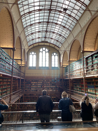 Old library in Rijksmuseum, Amsterdam. Tall windows with sunlight streaming through to old bookshelves. In foreground see backs of tourists as they view library from a balcony