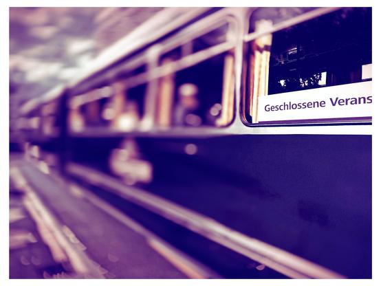 Das Bild zeigt einen Teil eines Zugwagens, der sich in Bewegung befindet. Der Zug hat eine dunkle Farbe, und die Fenster sind teilweise verschwommen, was auf eine schnelle Bewegung hinweist. Im Fenster ist ein Schild mit der Aufschrift "Geschlossene Verans" zu sehen, was auf Deutsch "geschlossene Anlage" bedeutet. Die Umgebung ist unscharf, was die Bewegung des Zuges betont. Die Farben des Bildes sind in einem violetten Ton gehalten, was eine nostalgische Atmosphäre vermittelt.