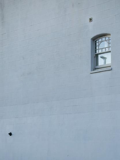 Photo of a whitewashed wall, the outlines of bricks can just be made out through the paint. In the top right is a small gently arched sash window, the top half split into a semi-circle pane surrounded by smaller ones arranged in a grid. The lamp atop a post is reflected in the bottom half. Above the window is what might be a small vent, dark disc within a black square. At the bottom left is a small black fixture, like a beauty mark.