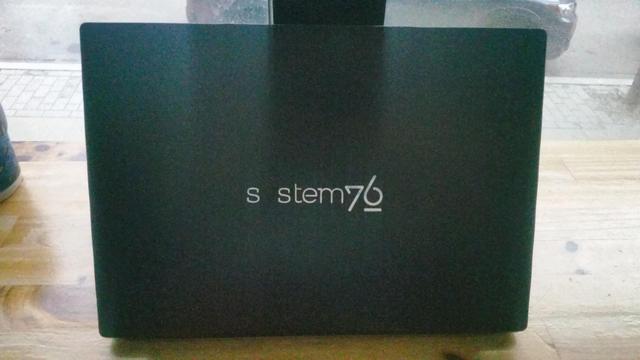 The back of a laptop, it says "s stem76" in the system76 logo style.

It's on a wooden table pointing away from a window.