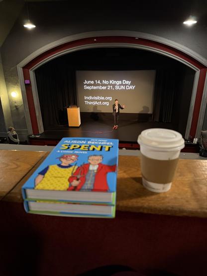 A photo taken from the balcony of the Brattle Theater in Cambridge, MA. In the foreground are two copies of Alison Bechdel's new book "Spent" and a cup of rather nice tea. In the background is Alison Bechdel herself talking to the audience from the stage. The projected slide behind her reads:
  June 14, No Kings Day
  September 21, SUN DAY

  Indivisible.org
  ThirdAct.org