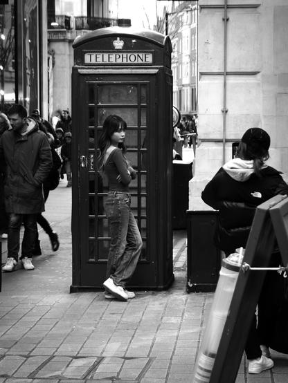 Black and white. A dark-haired young woman wearing jeans and a long-sleeved t-shirt leans against the door of a London telephone box while her friend takes a photo with her phone.