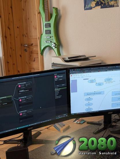 A desk with two screens. The screen on the left shows a node-based editor in dark mode and the screen on the right shows a diagram. A green guitar hangs on the wall in the background.