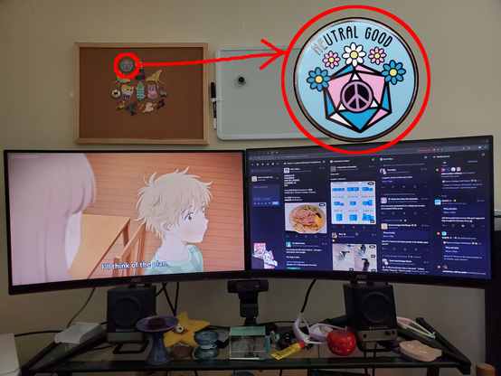 A photo of Kali Ranya's desktop arrangement. It has two side-by-side, landscape oriented monitors. Above the desk is a corkboard with various pins affixed. One is zoomed in with a scribbly callout: an emblem of a twenty-sided die, peace symbol, and flowers in transgender pride colors, labeled "Neutral Good."