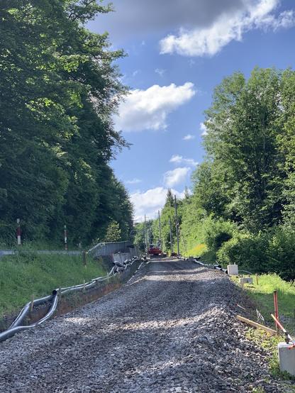 Blick bergwärts. Ein einsamer Bagger steht in einiger Entfernung auf den Schotterbett. Links und rechts hohe Bäume, der Himmel mit grossen Wolken, aber sonnig.