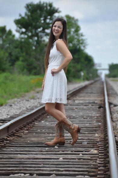 Emily Grice in white dress to her knee with cowboy boots. Long brunette hair. Standing in the middle of railroad tracks posing for graduation photos. Left hand on hip, looking left.