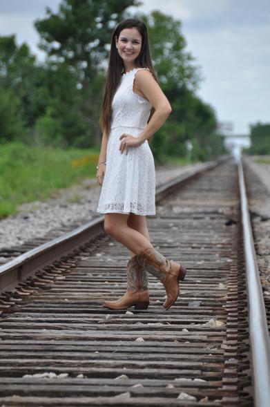 Emily Grice in white dress to her knee with cowboy boots. Long brunette hair. Standing in the middle of railroad tracks posing for graduation photos.